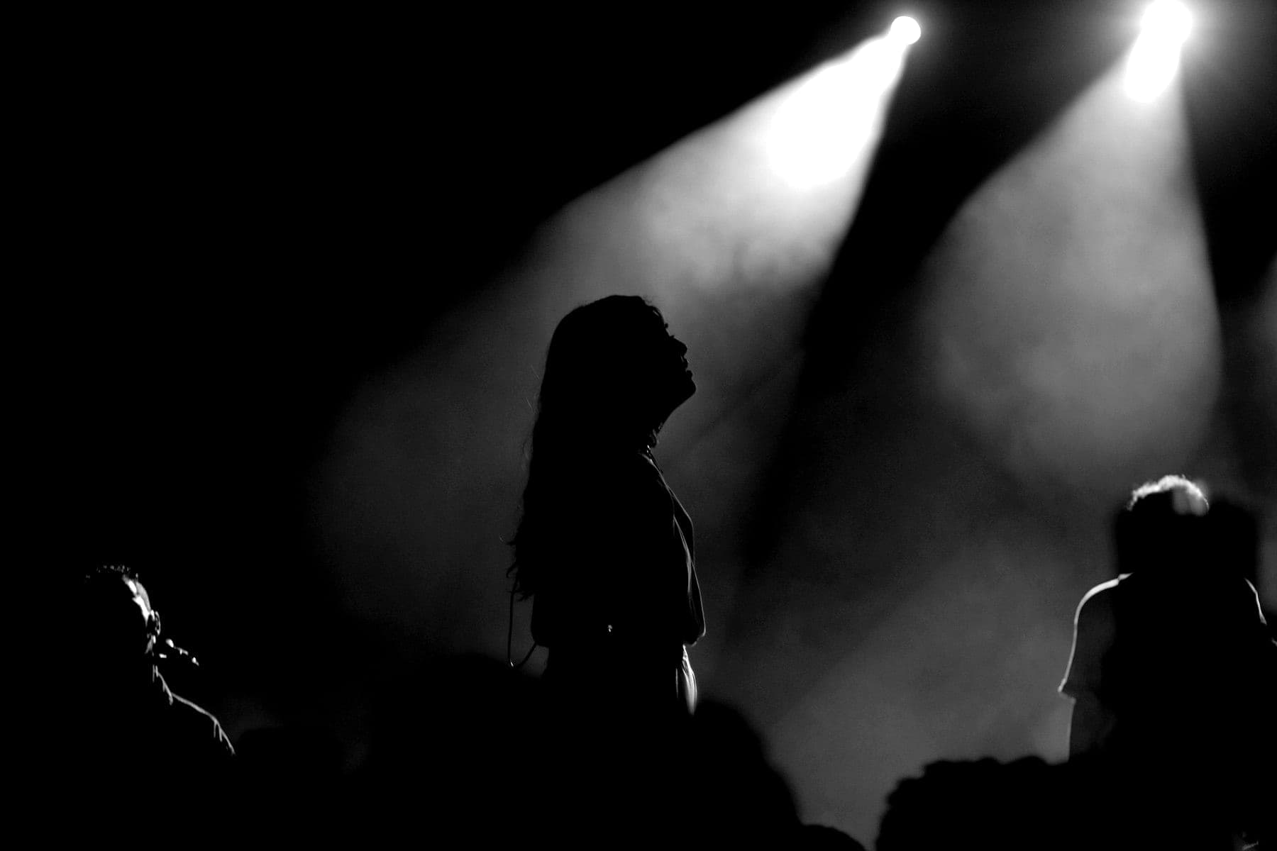 A performer in silhouette on stage, looking up into two spotlights in the haze.