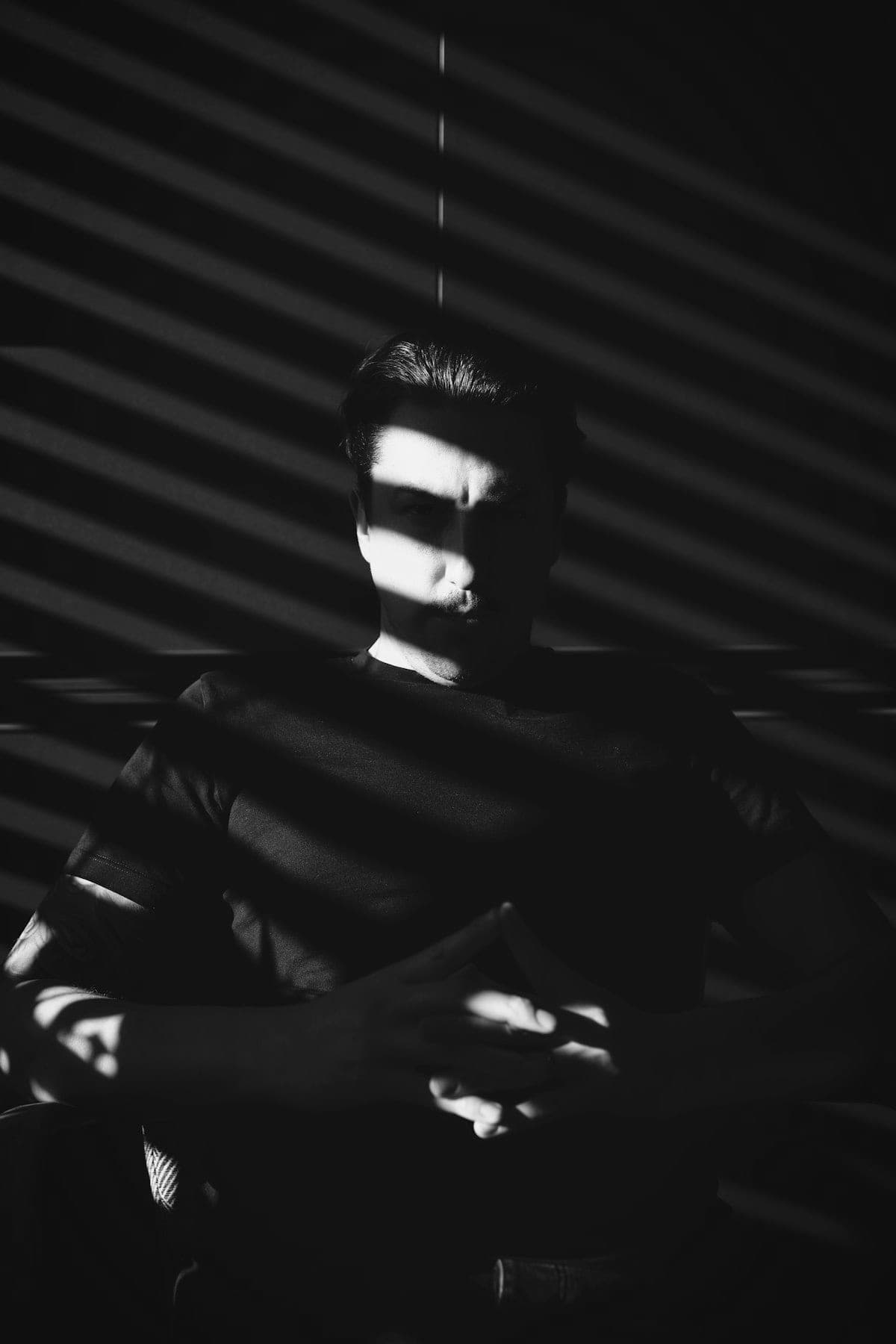 A man in a black T-shirt, face and body striped by hard shadow from window blinds.