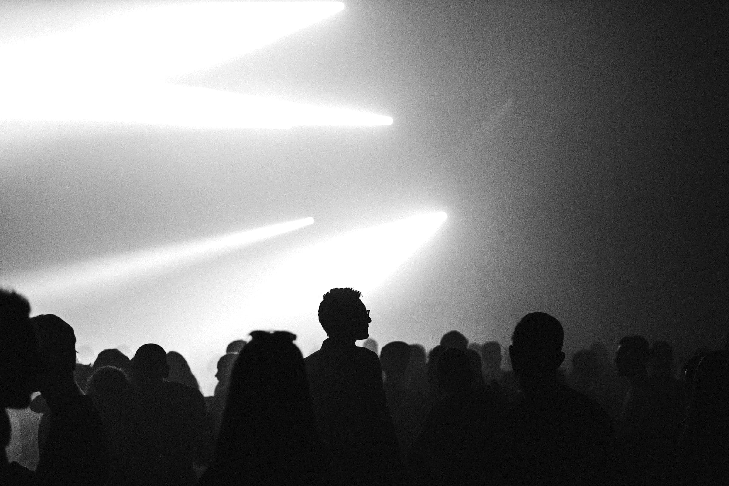 A concert crowd in silhouette beneath bright beams of stage light.