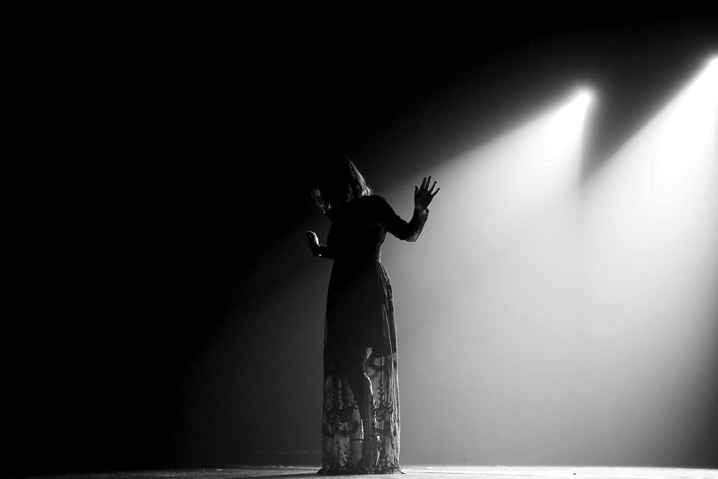 A performer in a long lace dress stands on a dark stage, one hand raised towards two spotlights cutting through the haze.