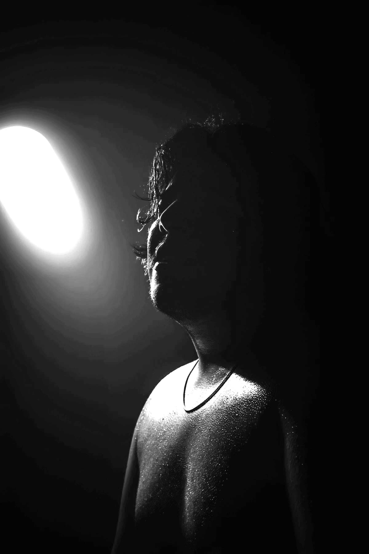 Black-and-white portrait: a figure in darkness, face mostly in shadow, turned towards a single spotlight.